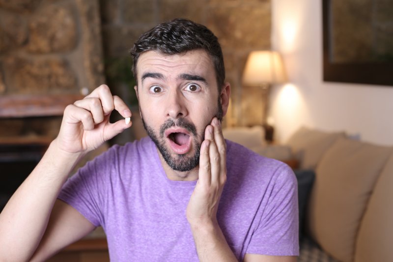 Shocked man in purple shirt holding his cheek in one hand and a lost tooth in another